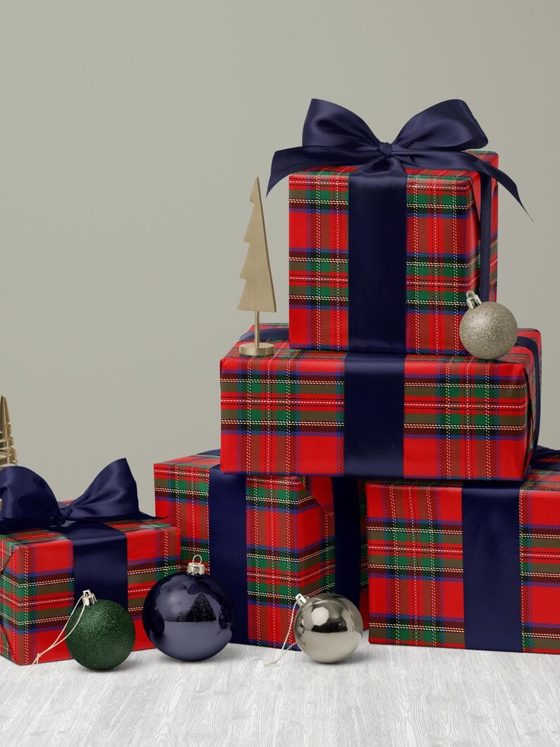 May include: Christmas-themed gift boxes wrapped in red plaid paper with navy blue ribbon and bows. Gold and silver ornaments and a gold tree decoration are arranged on a white wooden surface.