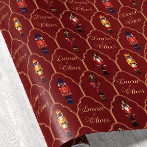 May include: Roll of burgundy wrapping paper with a repeating pattern of toy nutcrackers in red, gold, and black. Each nutcracker is set within a gold-lined, scalloped frame, with the text "Laura & Oliver" in elegant script.