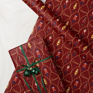 May include: Burgundy wrapping paper with a repeating pattern of nutcracker figures and the name "Emma" in gold script. A wrapped gift with green ribbon and ornaments is also visible.