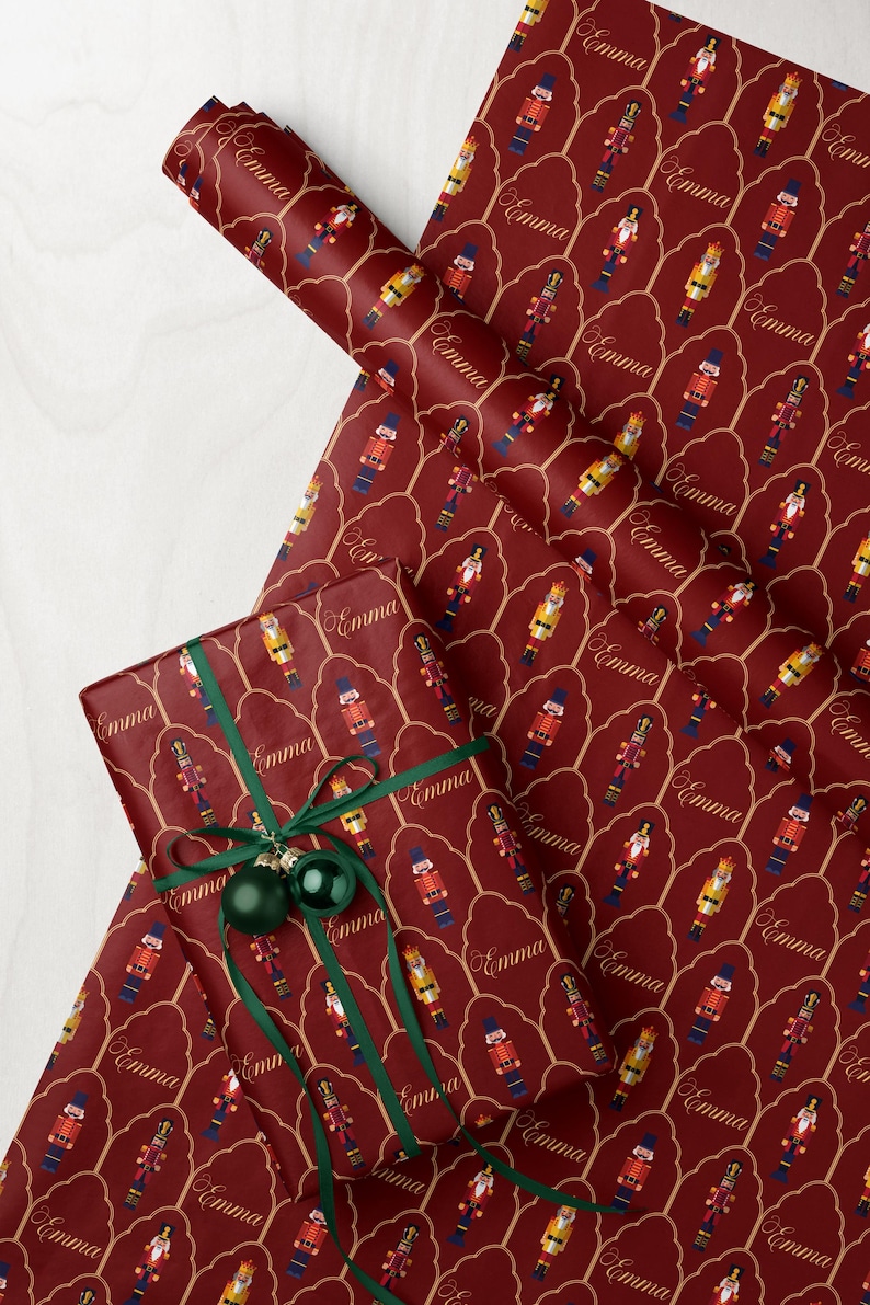May include: Burgundy wrapping paper with a repeating pattern of nutcracker figures and the name "Emma" in gold script. A wrapped gift with green ribbon and ornaments is also visible.