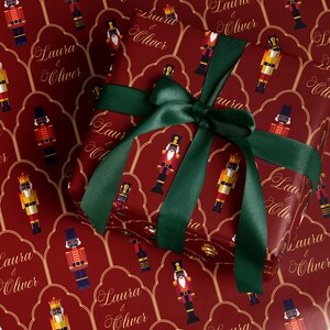 May include: A wrapped present with red wrapping paper patterned with colourful nutcracker figures. The gift is tied with a dark green satin ribbon and bow. The wrapping paper also features the text "Laura & Oliver" in gold script.