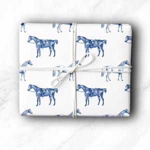 May include: White gift wrap featuring a repeating pattern of blue horse illustrations. The wrapped package is tied with white twine. The horses are depicted with saddles and bridles, creating a classic equestrian theme.