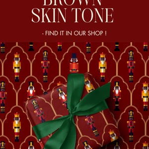 May include: A wrapped gift with a green ribbon sits on a red background patterned with nutcracker figures. The wrapping paper features various nutcracker designs in red, yellow, black, and blue. Text at the top reads "ALSO AVAILABLE WITH BROWN SKIN TONE - FIND IT IN OUR SHOP!"