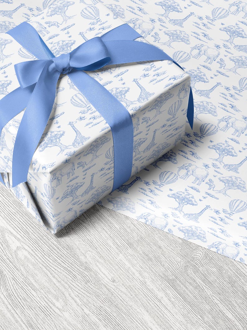 May include: A wrapped gift box with a light blue satin ribbon bow. The gift wrap features a white background with a blue safari animal pattern, including giraffes, elephants, and hot air balloons. The gift sits on a light wood grain surface.