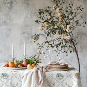 May include: A table setting featuring a white tablecloth with a repeating book and wreath pattern, two white taper candles, a bowl of assorted apples, two clear wine glasses, a stack of ceramic plates, and a tall branch with peach roses.