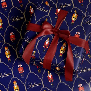 May include: A wrapped gift with a dark blue background featuring a repeating pattern of nutcracker soldiers and the name "Adam" in gold script. The gift is tied with a deep red satin ribbon and bow, creating a festive holiday presentation.