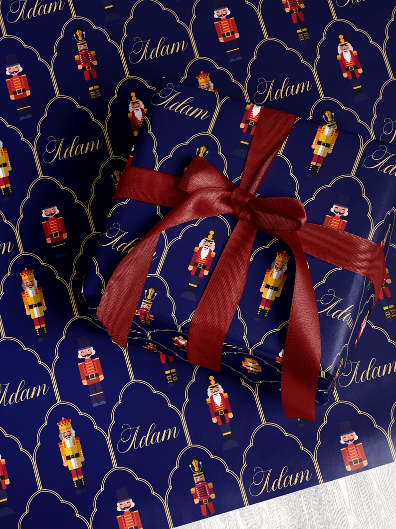 May include: A wrapped gift with a dark blue background featuring a repeating pattern of nutcracker soldiers and the name "Adam" in gold script. The gift is tied with a deep red satin ribbon and bow, creating a festive holiday presentation.