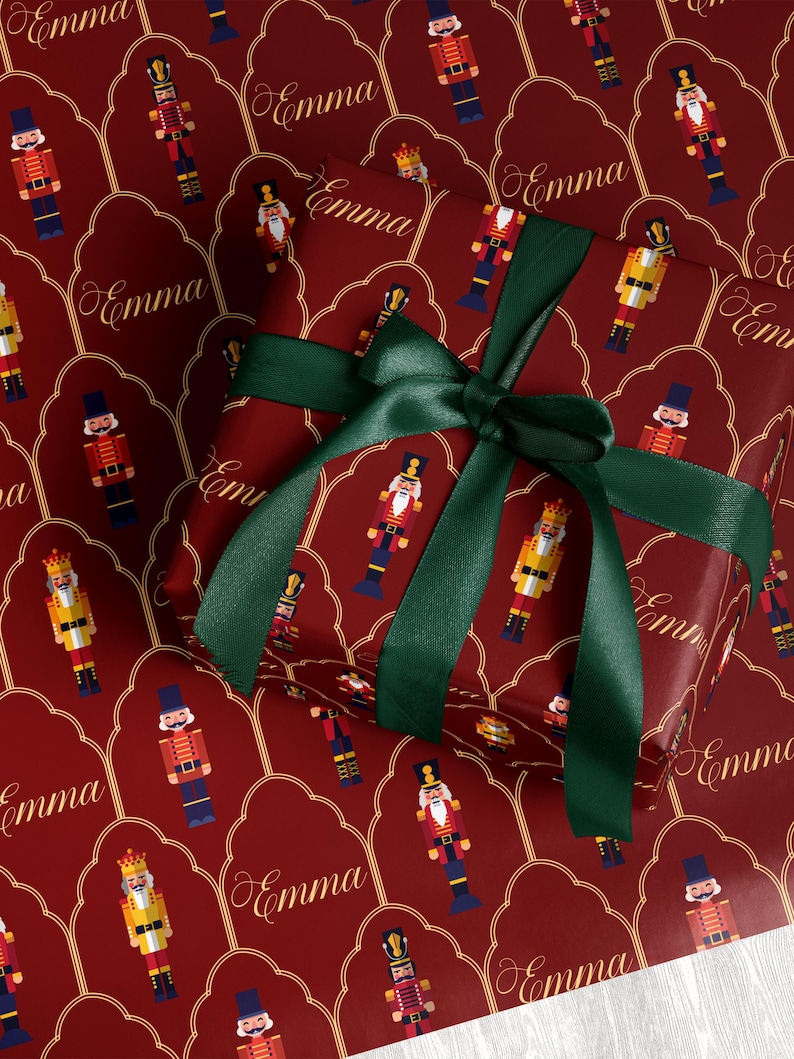 May include: A wrapped gift with a dark red background featuring a pattern of nutcracker soldiers and the name "Emma" in gold script. The gift is tied with a dark green satin ribbon and bow.