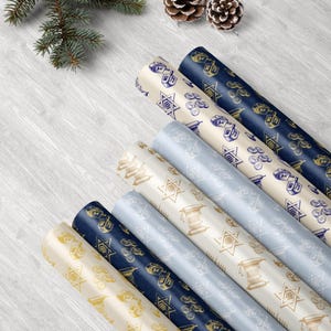 Puede incluir: Varios rollos de papel de regalo estampados en crema, azul marino y azul claro, con s&iacute;mbolos jud&iacute;os. El papel est&aacute; decorado con ilustraciones doradas y azul marino de dreidels, estrellas de David y otros motivos festivos. Una ramita de hoja perenne y pi&ntilde;as est&aacute;n en el fondo.