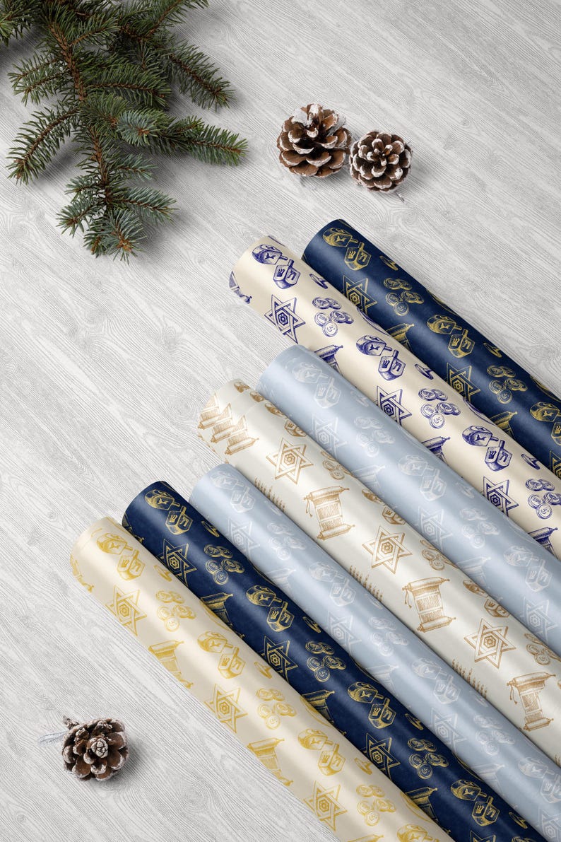 Puede incluir: Varios rollos de papel de regalo estampados en crema, azul marino y azul claro, con s&iacute;mbolos jud&iacute;os. El papel est&aacute; decorado con ilustraciones doradas y azul marino de dreidels, estrellas de David y otros motivos festivos. Una ramita de hoja perenne y pi&ntilde;as est&aacute;n en el fondo.
