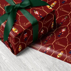 May include: A wrapped gift with a dark red background featuring a pattern of toy soldiers and the name "Emma" in gold script. The gift is tied with a dark green ribbon and bow.