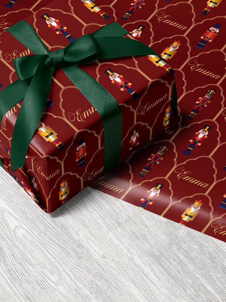 May include: A wrapped gift with a dark red background featuring a pattern of toy soldiers and the name "Emma" in gold script. The gift is tied with a dark green ribbon and bow.