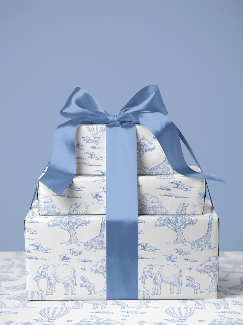 May include: Three stacked gift boxes wrapped in white paper with a blue safari animal print, tied with a large blue satin bow. The print features elephants, giraffes, trees, and hot air balloons.