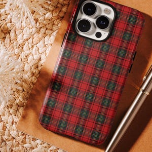 May include: A red and green plaid phone case with a black border. The case is on a brown surface with a woven texture. A silver pen is in the background.