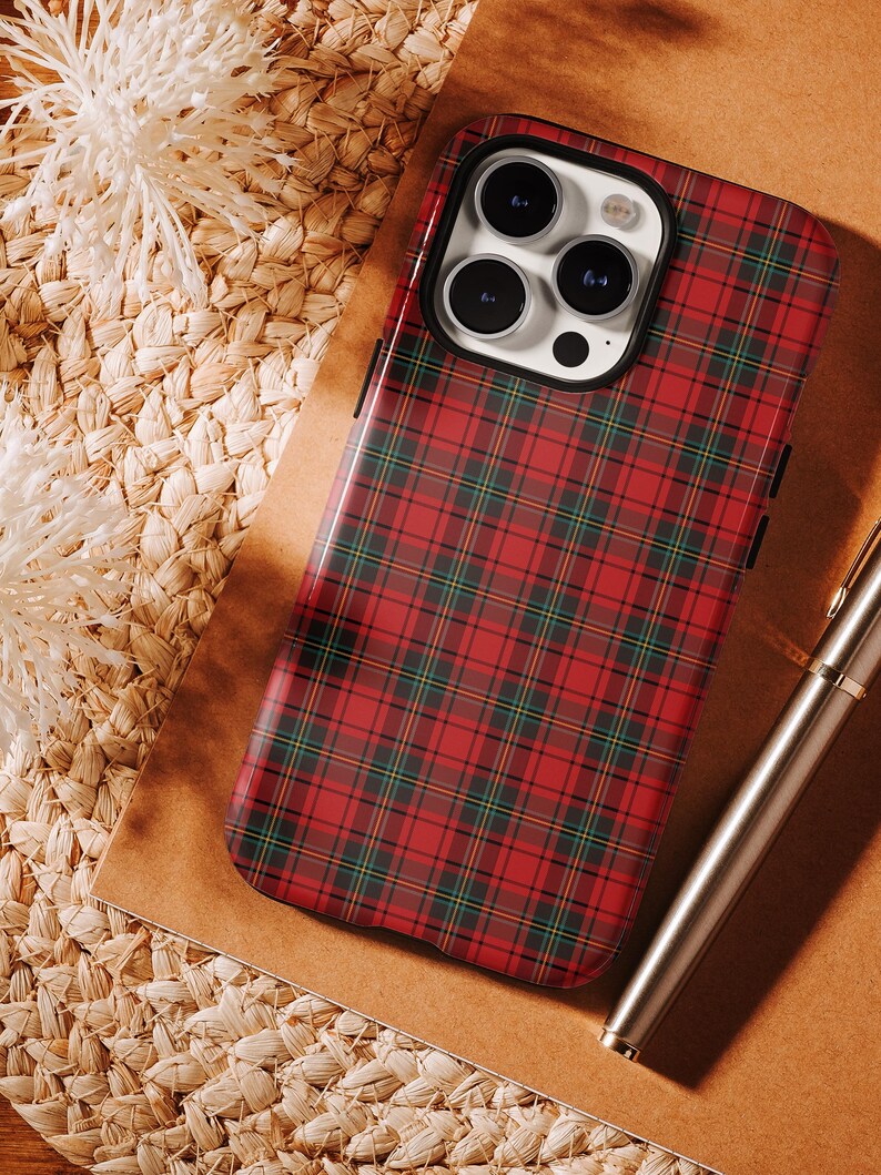 May include: A red and green plaid phone case with a black border. The case is on a brown surface with a woven texture. A silver pen is in the background.