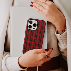May include: A red and green plaid phone case is held in front of a white book. The phone case has a black border and a camera cutout. The person holding the phone case is wearing a white coat and a sweater.