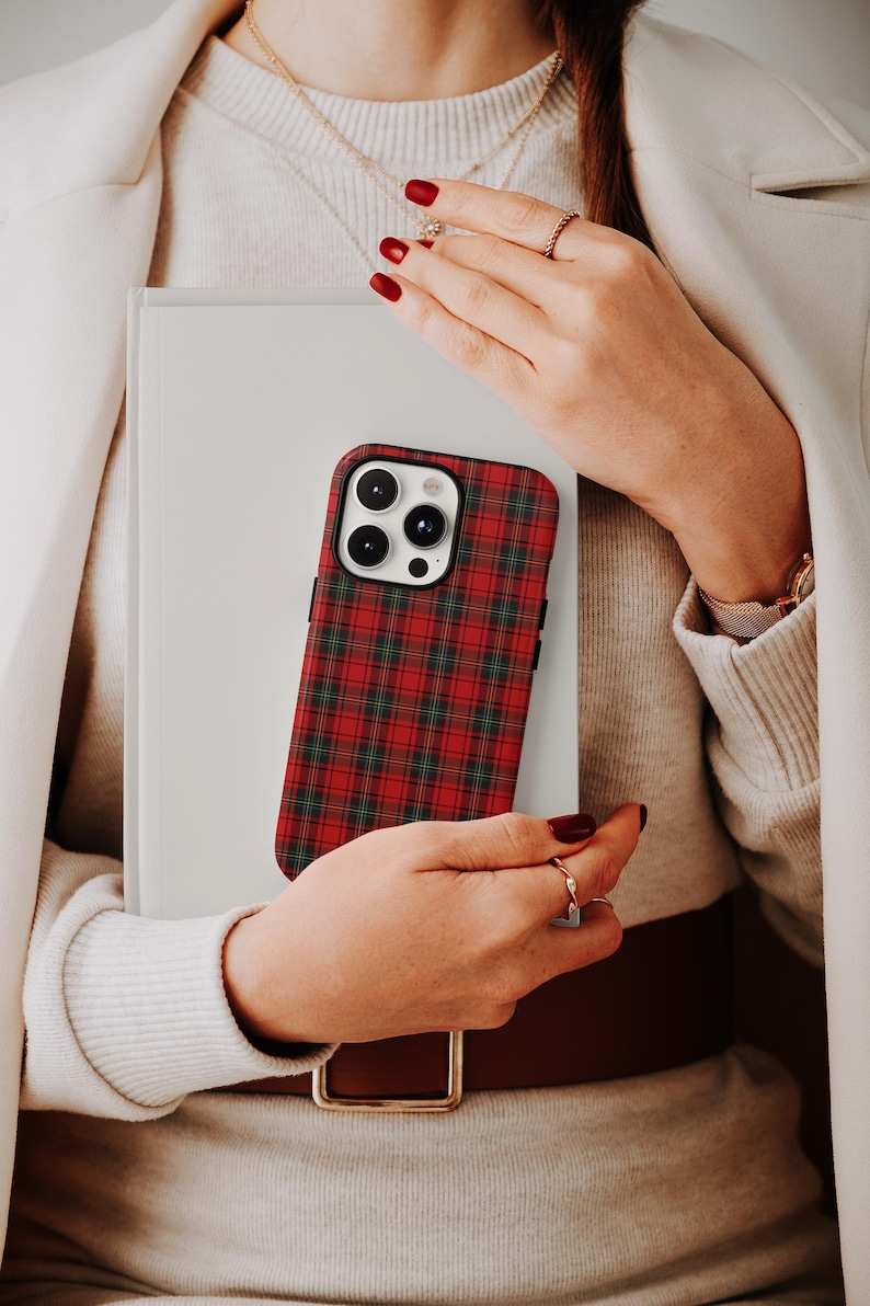 May include: A red and green plaid phone case is held in front of a white book. The phone case has a black border and a camera cutout. The person holding the phone case is wearing a white coat and a sweater.