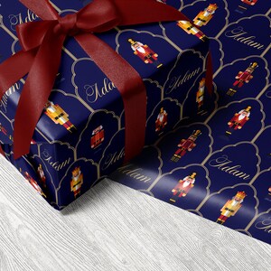 May include: A wrapped gift with a dark blue background featuring a pattern of nutcracker figures and the name "Adam" in gold script. The gift is tied with a deep red satin ribbon and bow.
