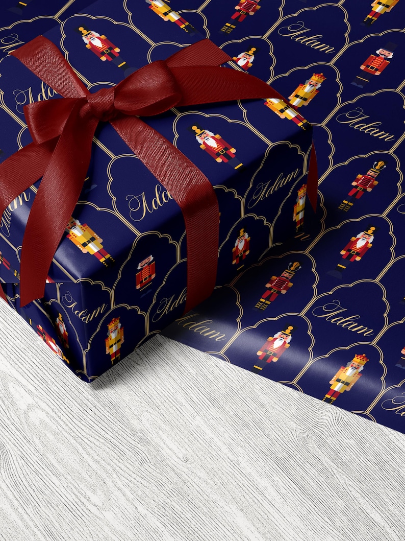 May include: A wrapped gift with a dark blue background featuring a pattern of nutcracker figures and the name "Adam" in gold script. The gift is tied with a deep red satin ribbon and bow.