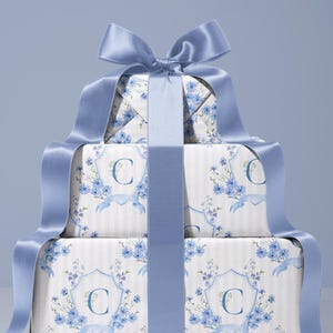 May include: Three gift boxes, wrapped in white paper with blue floral and stripe patterns. Each box features a large blue letter "C" and is tied with a wide, light blue satin ribbon and bow.