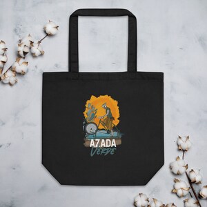 May include: Black tote bag with a graphic of a person riding a bicycle with the text "AZADA VERDE" in white and teal. The illustration features an orange sun and blue water elements.