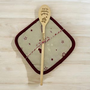 Könnte beinhalten: Ein Holzlöffel mit eingraviertem Text und Lebkuchenmann-Design, der auf einem hellgrünen Topflappen mit bordeauxrotem Rand und Sternmustern liegt. Löffel und Topflappen sind mit rot-weißem Garn zusammengebunden.