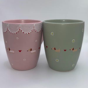 Könnte beinhalten: Zwei Keramikbecher, einer rosa und einer grün, mit Dackel- und Herzmotiven. Der rosa Becher hat einen weißen, gewellten Rand mit roten Punkten. Der grüne Becher hat weiße Gänseblümchen-Akzente. Beide Becher haben eine glatte, glänzende Oberfläche.