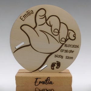 Könnte beinhalten: Eine Holzplakette mit einem Handabdruck-Design und dem Namen "Emilia" eingraviert. Die Plakette enthält auch das Datum "16.01.2024", die Uhrzeit "07:36 Uhr", das Gewicht "3420g" und die Länge "52cm".
