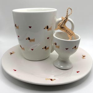 Könnte beinhalten: Ein Frühstücksset aus weißer Keramik bestehend aus einer Tasse, einem Eierbecher und einem Teller, verziert mit einem Muster aus kleinen braunen Dackeln und winzigen roten Herzen. Ein Dackel-förmiger Holzanhänger ist am Henkel der Tasse befestigt.