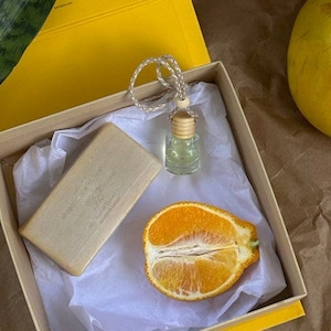 May include: A yellow gift box with a white tissue paper lining. The box contains a bar of soap, a small bottle of essential oil, and a halved orange. A green plant and a yellow grapefruit are visible outside the box.