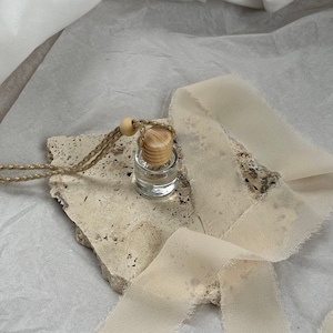 Könnte beinhalten: Eine kleine Glasflasche mit einem Holzverschluss und einem beigefarbenen Band, das um sie herum gebunden ist. Die Flasche steht auf einem Stück beigefarbenem Stein mit rauer Textur.