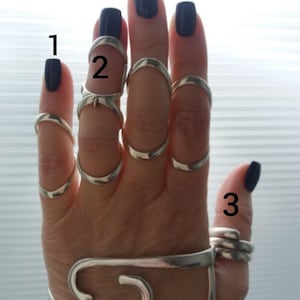 May include: A hand wearing multiple silver rings, including knuckle rings and a bracelet-like piece. The rings are of various designs, some covering entire knuckles. The nails are painted a dark blue color.