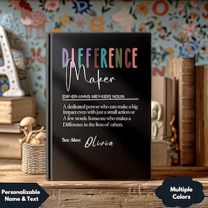 May include: A black notebook with the words "DIFFERENCE Maker" in colorful text. The definition of "Difference Maker" is written below, followed by "See Also: Olivia". The notebook is displayed on a wooden surface.
