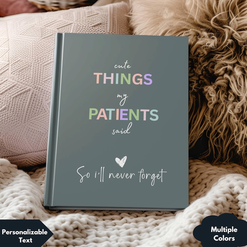 May include: A dark gray journal with the text 'cute THINGS my PATIENTS said So I'll never forget' in rainbow colors. The text is in a handwritten font. There is a small heart icon above the last line of text. The journal is sitting on a white fuzzy surface.