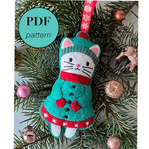 May include: A white felt cat ornament with a red scarf and green coat, wearing a red and white striped hat. The ornament is hanging on a green pine branch with other Christmas decorations.