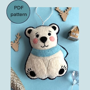May include: A handmade white felt polar bear ornament with a blue scarf and black accents. The bear has pink cheeks and black paws. The ornament is part of a PDF pattern, as indicated by the text in a blue circle. Other decorative elements include wooden reindeer and a small house.