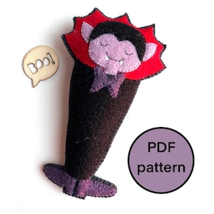 May include: A felt vampire doll with a red collar and purple details. The vampire has a smiling face and a purple bow tie. A wooden speech bubble with "Boo!" is next to the doll. A purple circle with the text "PDF pattern" is also in the image.