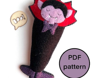May include: A handmade felt vampire doll with a red collar and purple accents. The vampire has a smiling face and is wearing a purple bow tie. A wooden speech bubble with the word "Boo!" is next to the doll. A purple circle with the text "PDF pattern" is also in the image.