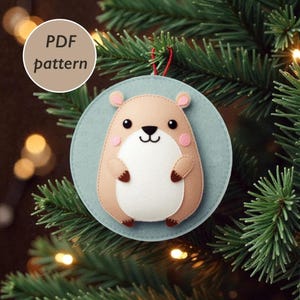 Könnte beinhalten: Handgefertigter Weihnachtsbaumschmuck aus Filz in Form eines Hamsters, mit hellbraunem Körper, weißem Bauch und rosa Wangen. Der Schmuck ist an einem hellblauen Kreis befestigt und hat eine rote Schnur zum Aufhängen. Der Text "PDF pattern" ist in einem Kreis.