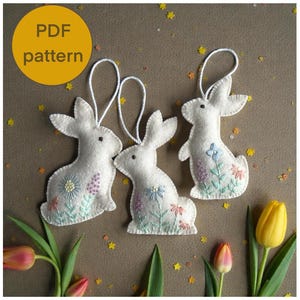 Könnte beinhalten: Drei weiße Hasen-Ornamente aus Filz mit gestickten Blumenmustern in Pastellfarben. Jeder Hase hat eine weiße Schnurschlaufe zum Aufhängen. Das Bild enthält den Text "PDF pattern" in einem gelben Kreis.