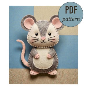 Könnte beinhalten: Eine handgefertigte Filzmaus mit grauen, weißen und rosa Details. Die Maus hat eine genähte Kontur, schwarze Schnurrhaare und einen rosa Schwanz. Ein blauer Kreis mit dem Text "PDF pattern" ist zu sehen.