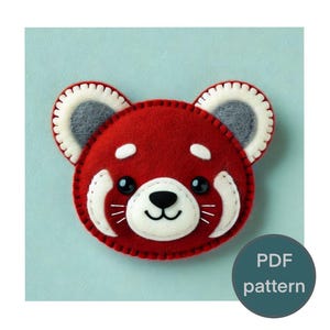 May include: A handmade red panda felt applique with white accents and black eyes. The panda has white markings around the eyes and mouth, with a black nose and a smiling mouth. The ears are gray with white trim. The text "PDF pattern" is in a teal circle.