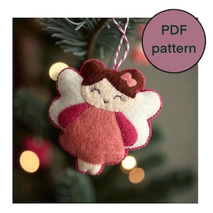 Könnte beinhalten: Handgefertigter Feen-Ornament aus Filz mit lächelndem Gesicht, braunen Haaren und einer rosa Schleife. Die Fee hat weiße Flügel und ein rosa Kleid. Das Ornament hängt an einer Schnur. Eine runde Grafik zeigt "PDF pattern".