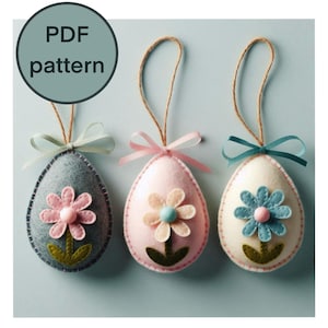 Op de afbeelding: Drie vilten paaseieren ornamenten met een bloemmotief. De eieren zijn in tinten roze, grijs en wit. Elk ei heeft een bloem in een andere kleur met een wit centrum en een groene steel.