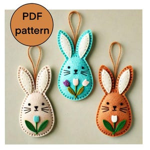 May include: Three felt Easter bunny decorations with a flower design. The bunnies are light brown, light blue, and orange. Each bunny has white felt ears and a white felt flower with a green stem.