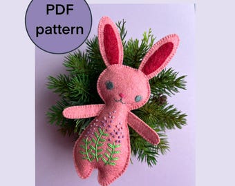 May include: Pink felt bunny with embroidered green leaves and purple flowers. The bunny has red felt ears and a stitched smile.