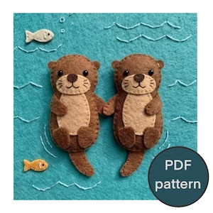 May include: Two brown felt otters holding hands, set against a teal background with felt fish and water lines. The otters have tan bellies and embroidered details. A circular graphic in the bottom right corner reads "PDF pattern".
