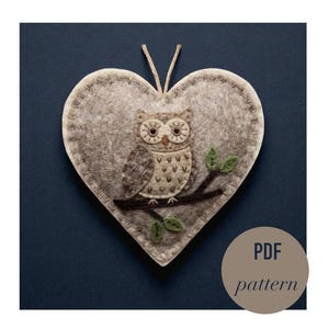 May include: A heart-shaped felt decoration featuring a stitched owl on a branch, with green leaves. The owl is in brown and beige tones. It has a beige string for hanging. The text "PDF pattern" is in a circle.