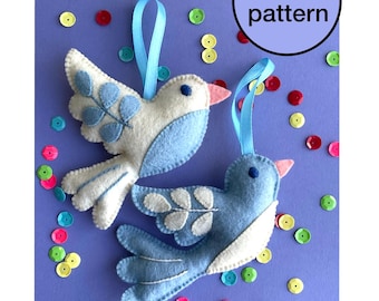 May include: Two felt bird ornaments in white and blue with pink beaks and blue ribbon hangers. The birds are decorated with felt leaves and are on a purple background scattered with colorful sequins. A circular graphic reads "PDF pattern".