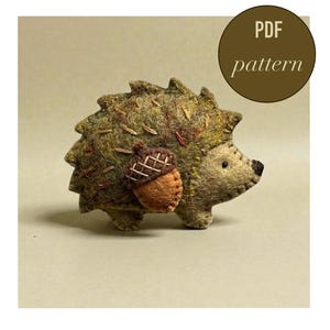 May include: A handmade felt hedgehog with a brown acorn attached. The hedgehog has a textured, green and brown body with embroidered details. The image includes a circular graphic that says "PDF pattern".